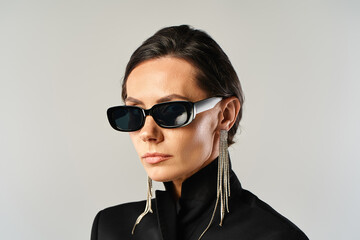 A fashionable woman with sunglasses poses in a black jacket against a grey studio backdrop.