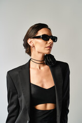 A fashionable woman exudes confidence in a black suit and sunglasses, striking a pose in a studio against a grey background.
