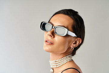 A stylish woman with sunglasses and a pearl necklace exudes confidence in a studio against a grey...