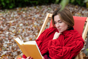 A woman sits on a bench on the backyard as she gets lost in the pages of her book. The autumn...
