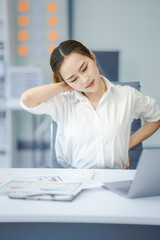 A young businesswoman sits at her desk in the office, stretching her tired muscles. She feels pain in her wrist and shoulder, suffering from the strain of hard work.