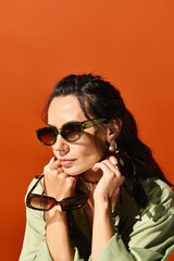 A stylish woman wearing sunglasses strikes a confident pose in a studio against a vibrant orange...