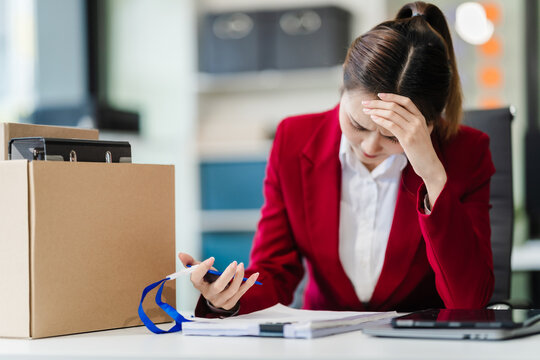 In the office, an Asian woman carefully signs her resignation letter, feeling stressed. She packs her belongings in cardboard boxes, preparing to leave the workplace. Asian people.