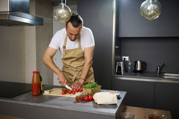 Handsome young man in beige chef's apron, cooking healthy vegetarian salad in the modern minimalist home kitchen, standing at kitchen island and chopping fresh ripe cherry tomatoes.
