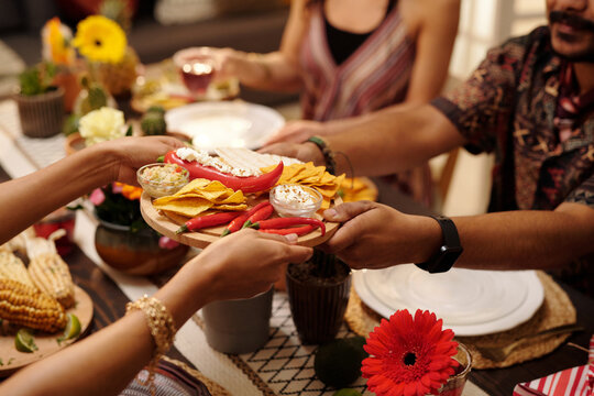 Hands of young woman passing tray with nachos, red hot chili peppers and savory sauce to male guest sitting by served festive table