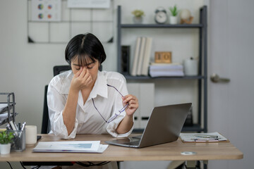 Exhausted female office worker feeling stressed, holding glasses, with laptop and documents on desk.