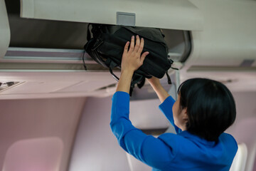 A traveler in a blue jacket is placing her carry-on bag into the overhead storage compartment on an airplane.