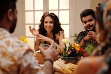 Young smiling brunette woman looking at one of friends during chat by festive table at home party while sitting next to Hispanic man