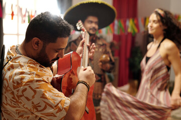 Young bearded male musician in national attire playing guitar in front of camera while dynamic man and woman dancing at home party