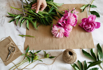 Fototapeta na wymiar Florist at work: pretty woman making summer bouquet of peonies on a working gray table. Kraft paper, scissors, envelope for congratulations on the table. View from above. Flat lay composition.
