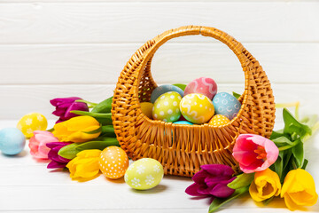 Easter basket filled with colorful eggs and a bouquet of tulips on a textured wooden table. Easter celebration concept. Colorful easter handmade decorated Easter eggs. Place for text. Copy space