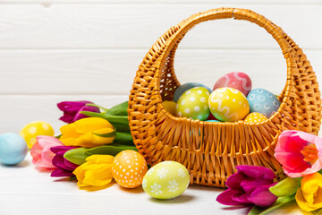 Easter basket filled with colorful eggs and a bouquet of tulips on a textured wooden table. Easter...