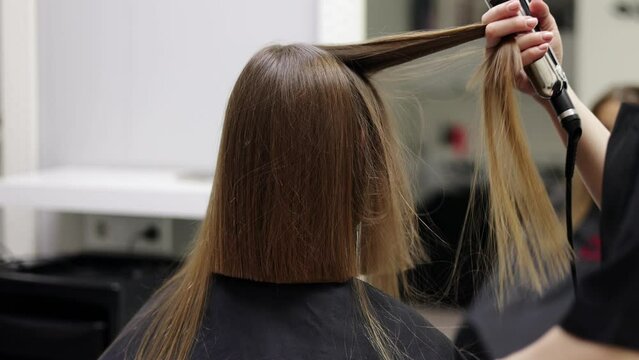 Unrecognisable hairdresser uses comb and hair styler for straighten hair for prepares to cuts female client hair. Professional barber makes stylish haircut short bob hairstyle in hair salon. care