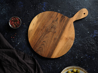 Top view of empty round wooden cutting board with handle, red pepper and green olives on abstract dark background.