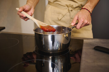 Close-up chef standing by electric induction stove, using wooden ladle, stirring boiling tomato...