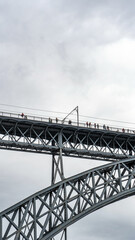 Tourists walking, taking photos and a woman posing from the upper platform of the Don Luis I steel bridge where the Porto metro passes with rain clouds.