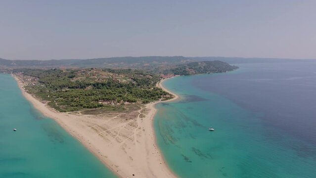 The drone moves backwards over Possidi Cape, Halkidiki, Greece, showing the sandbar, clear turquoise sea, and the surrounding forested area