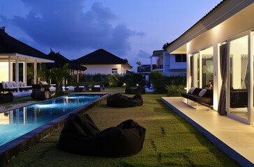Modern Bali-style house with pool and garden, night view, black bean bag chairs around the corner of an open-air area with green grass near the swimming pool, white walls with dark windows