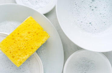 Yellow sponge and a soapy foam, white plate with soap suds on a background. Cleaning concept, cleaning service, flat lay, top view - 782225815