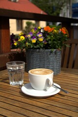 Cafe latte in a cup and glass of water on a table decorated with pansies in an ornamental flower pot
