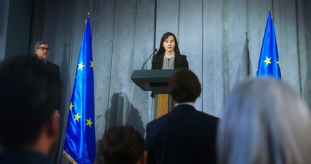 Confident politician makes an announcement, answers media questions and gives interview. Female representative of the European Union during performance at press conference. Backdrop with EU flags. - obrazy, fototapety, plakaty