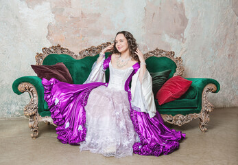 Smiling beautiful woman in fantasy white and purple rococo style medieval dress sitting on the sofa