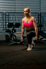 Fototapeta na wymiar in the gym a girl with a ponytail in a pink top does lunges with dumbbells in her hands