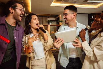 Multiracial group of friends having karaoke night. Singing in to the microphone, looking at lyrics and drinking beer. - obrazy, fototapety, plakaty
