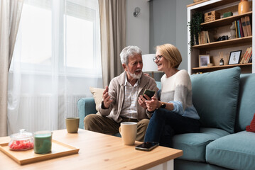 Senior couple, simple living. Older people man and woman full of love and respect, sitting at cozy home talking looking online funny videos on smartphone, enjoying their retirement and life.