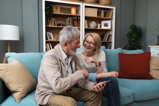Senior couple, simple living. Older people man and woman full of love and respect, sitting at cozy home talking looking online funny videos on smartphone, enjoying their retirement and life.