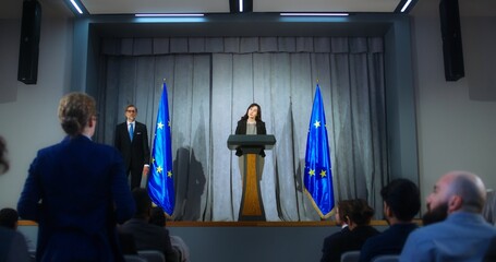 Female representative of the European Union during press conference. Confident politician makes an announcement, answers journalists questions and gives interview for media. Backdrop with EU flags. - obrazy, fototapety, plakaty
