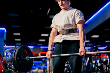 close up in the gym a young handsome guy in a T-shirt and a cap with a belt lifts a barbell with large athletic pancakes