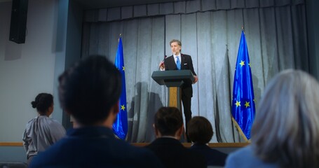 Mature representative of the European Union during performance at press conference. Confident politician makes an announcement, answers media questions and gives interview. Backdrop with EU flags. - obrazy, fototapety, plakaty