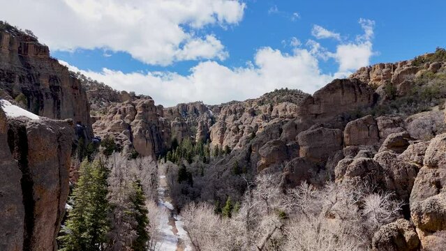 Aerial-High over snowy road along canyon lined with pine trees, bare winter trees and rugged geologic rock formations comprised of conglomerate material embedded in a sandstone base. 