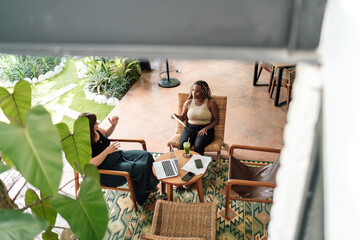 Two professional women, a Caucasian and an African American, engaged in a collaborative discussion over a work project on a patio with lush greenery.