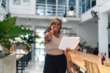 Young adult African-American female entrepreneur giving thumbs-up in a modern coworking space, holding documents, exuding confidence and professionalism.