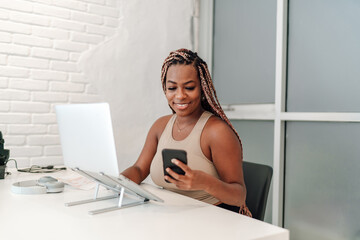 African-American woman entrepreneur in her 30s, business casual, working at white desk with a laptop computer while using smartphone application for managing tasks, cheerful in a well-lit office