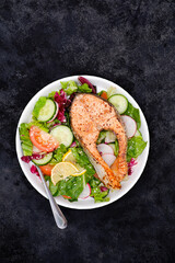 Grilled salmon and fresh vegetables: tomatoes, cucumber, lemon, radish on black background.