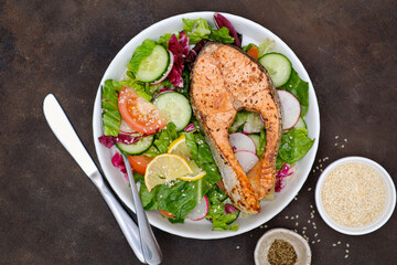 Grilled salmon and fresh vegetables: tomatoes, cucumber, lemon, radish on dark brown background.