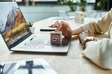 A modern businesswoman holds a small house model in front of a laptop, embodying innovation in...