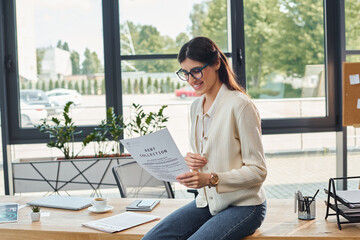 A modern businesswoman sits on a table in an office, engrossed in a book.