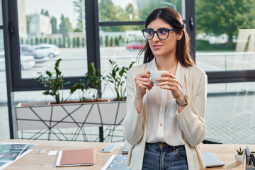 A sophisticated businesswoman in glasses cherishes a warm cup of coffee in a modern office,...
