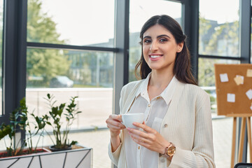 A businesswoman savors a cup of coffee against a cityscape backdrop through a large window in a...