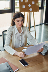 A modern businesswoman sits at a table, enjoying a moment of relaxation with a cup of coffee in a...