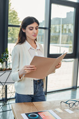 A businesswoman stands in a modern office, holding a piece of paper, with a table in the background...