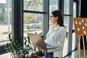 A modern businesswoman stands by a window, holding a laptop in her hands as she works on franchise...