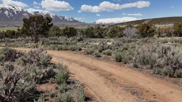 Aerial-Flying low over country dirt road and sagebrush hill dotted with juniper trees to view small rural mountain valley town in the distance.