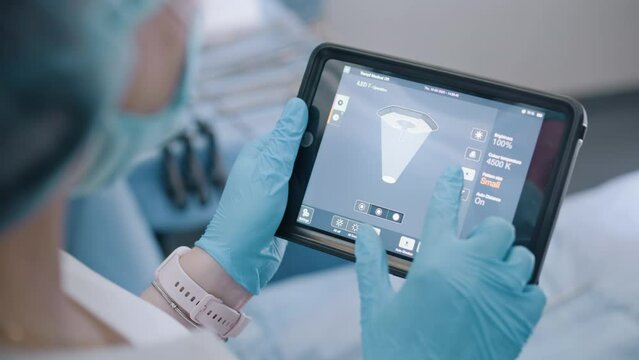 A surgical assistant adjusts the light on a lighting fixture in the operating room before starting a surgical operation using a digital tablet.