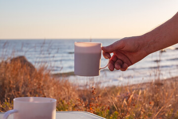 A mug with morning coffee in a man hand against the backdrop of the sea coast. Outdoor recreation, travel and tourism