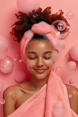 Happy and relaxed young woman after a shower or beauty treatment, with a pink towel on her head and soap bubbles on a pink background. Body care, beauty and relaxation treatments.
